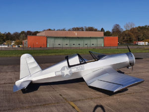 Photo of the ultralight Corsair replica on the runway in front of a colorful hangar building.